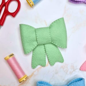 Può includere: Un fiocco in feltro verde chiaro con un design cucito. Il fiocco è circondato da rocchetti di filo di vari colori, tra cui rosa, blu e giallo, e un paio di forbici rosse. L'oggetto è un articolo artigianale.