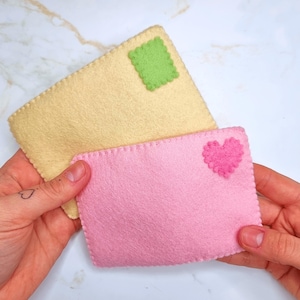 May include: Two felt envelopes, one pale yellow with a green square and one pink with a pink heart. Both are rectangular with stitched edges. The envelopes are held up against a white marble surface.