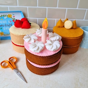 Puede incluir: Tres pasteles redondos de fieltro. El pastel delantero es rosa con remolinos de glaseado blanco y una vela. El pastel izquierdo es de color crema con tres fresas de fieltro rojas. El pastel derecho es marrón dorado con tres adornos amarillos.
