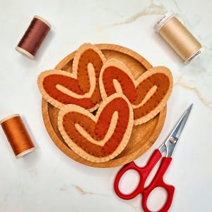 May include: A wooden plate holds heart-shaped felt decorations in shades of brown and orange. Three spools of thread in similar colors and a pair of red-handled scissors are arranged around the plate, suggesting a craft or sewing project.