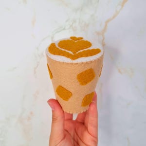 Puede incluir: Una taza de caf&eacute; de fieltro con una parte superior de espuma blanca y un dise&ntilde;o de coraz&oacute;n. La taza es marr&oacute;n con manchas amarillas.