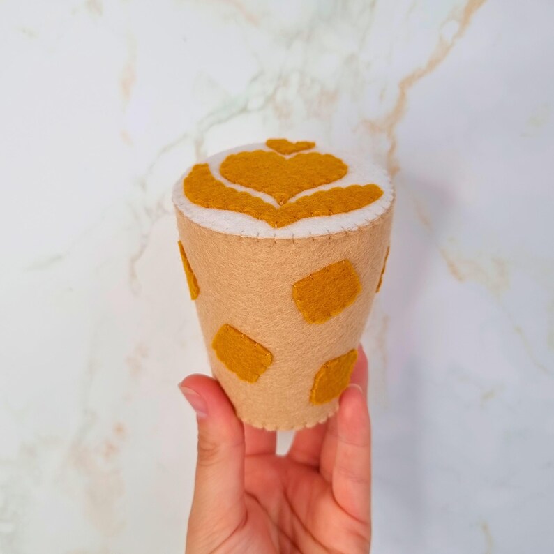 Puede incluir: Una taza de caf&eacute; de fieltro con una parte superior de espuma blanca y un dise&ntilde;o de coraz&oacute;n. La taza es marr&oacute;n con manchas amarillas.
