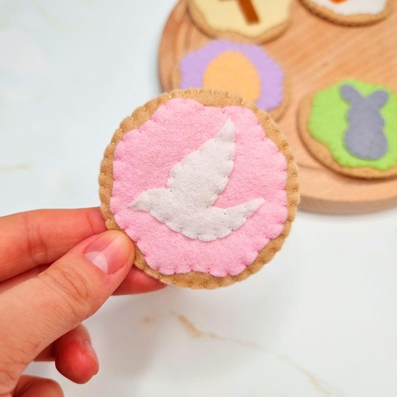 May include: A felt Easter cookie with a pink icing base and a white felt dove on top. The cookie is surrounded by a brown felt border.
