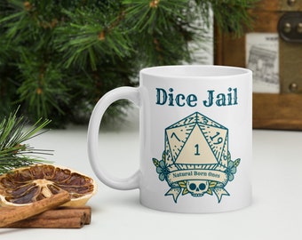 May include: White ceramic mug with the text "Dice Jail" and a graphic of a D20 die with the number 1. The design includes floral accents, a banner that reads "Natural Born Ones," and a skull. The mug has a curved handle.