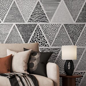 May include: A living room scene featuring a beige sofa with several throw pillows in shades of brown and black. A black and white patterned throw blanket is draped over the sofa. The wall behind the sofa is decorated with a black and white triangle pattern.