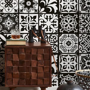 May include: A room scene featuring a black and white patterned tile wall. A wooden chair with a woven leather seat and a green blanket is on the left. A brown wooden nightstand with a textured front holds a glass, books, and binoculars.