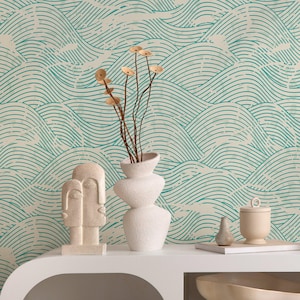 Op de afbeelding: Een witte plank toont decoratieve items tegen een turquoise golfpatroon behang. Items zijn onder meer een witte vaas met gedroogde stelen, een stenen gezichtssculptuur, een kleine klok en een decoratieve kom.