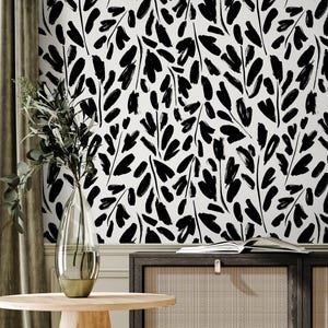 May include: A room interior featuring a black and white leaf pattern wallpaper. A wooden side table with a vase of greenery sits in front of a dark wood cabinet with woven panels. A cream-colored armchair and a patterned throw complete the scene.