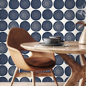 May include: A dining room scene featuring a round wooden table and two brown leather chairs. The background is a cream-colored wallpaper with a repeating pattern of navy blue concentric circles. A vase with dried flowers and tableware are on the table.