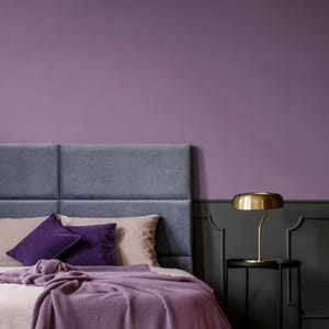 Könnte beinhalten: Schlafzimmer-Interieur mit einem Bett mit grauem Kopfteil, hellrosa Bettwäsche und lila Kissen und Decke. Eine goldfarbene Lampe steht auf einem schwarzen Beistelltisch. Die Wände sind in Lila- und Grautönen gestrichen.