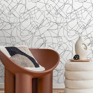 Può includere: Una scena d'interni moderna con una sedia marrone, un cuscino fantasia e un pouf impilato. Un vaso decorativo è posto sopra il pouf. Lo sfondo è una parete bianca con un motivo a scarabocchi neri. Pantofole in pelle nera sul tappeto.