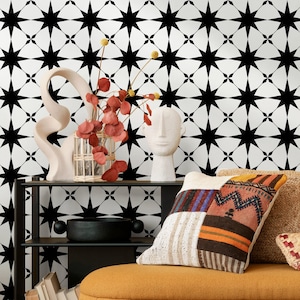 Op de afbeelding: Een decoratieve interieurscène met een zwart-wit sterrenpatroon behang. Een zwarte plank bevat een witte sculptuur, een vaas met gedroogde bloemen en een witte buste. Een patchwork kussen ligt op een gele bank.