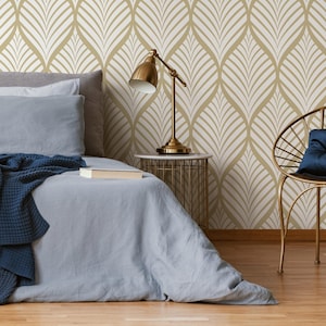 May include: A bedroom with a bed, a nightstand, a lamp, and a chair. The walls are covered in a gold and white geometric patterned wallpaper. The bed has a blue blanket and a white pillow. The nightstand is made of gold metal and has a white marble top. The lamp is gold and has a white shade. The chair is gold and has a white cushion.