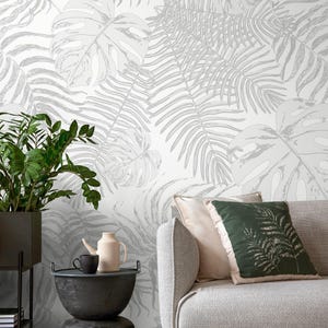 Puede incluir: Un salón con un sofá gris, cojines y papel pintado con un diseño de hojas tropicales en blanco y gris. Una mesa auxiliar negra con una planta, una tetera y una taza. Tema botánico moderno.