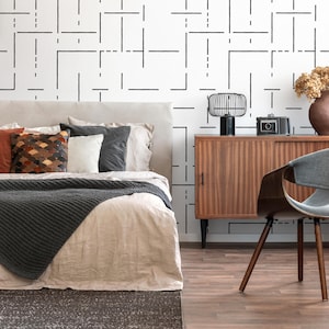 May include: A bedroom with a bed with a gray knitted throw blanket and white linen bedding. The bed has a headboard with several pillows. A wooden dresser with a black lamp and a camera on top is to the right of the bed. A gray chair with a curved back is in the foreground. The wall behind the bed has a black and white geometric pattern.