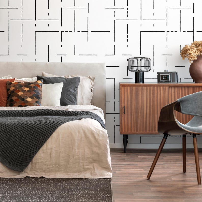 May include: A bedroom with a bed with a gray knitted throw blanket and white linen bedding. The bed has a headboard with several pillows. A wooden dresser with a black lamp and a camera on top is to the right of the bed. A gray chair with a curved back is in the foreground. The wall behind the bed has a black and white geometric pattern.