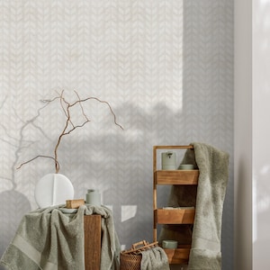 May include: A serene bathroom scene featuring a wooden shelf and a small wooden table. The shelf holds towels, soap, and a candle. A white vase with a branch sits on the table, draped with a green towel. The wall has a light gray chevron pattern.