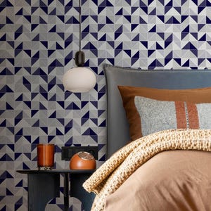 Puede incluir: Un dormitorio con papel pintado geométrico en azul marino, gris y blanco. Una mesita de noche con una vela, un despertador retro y una lámpara colgante. La cama tiene ropa de cama y almohadas marrones.