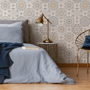 May include: A bedroom interior featuring a bed, a side table, a lamp, and a chair. The walls are adorned with patterned wallpaper featuring blue and gold geometric designs. The bed has a blue blanket and a white pillow. The side table is made of gold metal and has a white marble top. The lamp is also made of gold metal and has a white shade. The chair is made of gold metal and has a blue cushion.