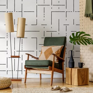 May include: A green armchair with a brown and white patterned pillow sits in a room with a white wall featuring a black geometric pattern. Two floor lamps with brown shades stand on either side of the chair. A small wooden side table with a black vase and a brown mug sits next to the chair.