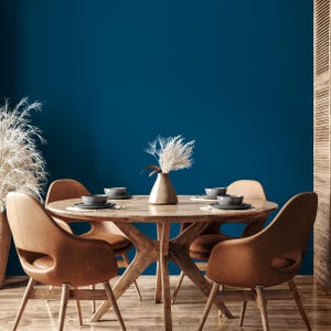 Puede incluir: Un comedor con una mesa redonda de madera y cuatro sillas marrones. La mesa está puesta con cuencos y platos grises, y un jarrón con flores secas está en el centro. El fondo es una pared azul oscuro. La mesa tiene unos 120 cm de diámetro.