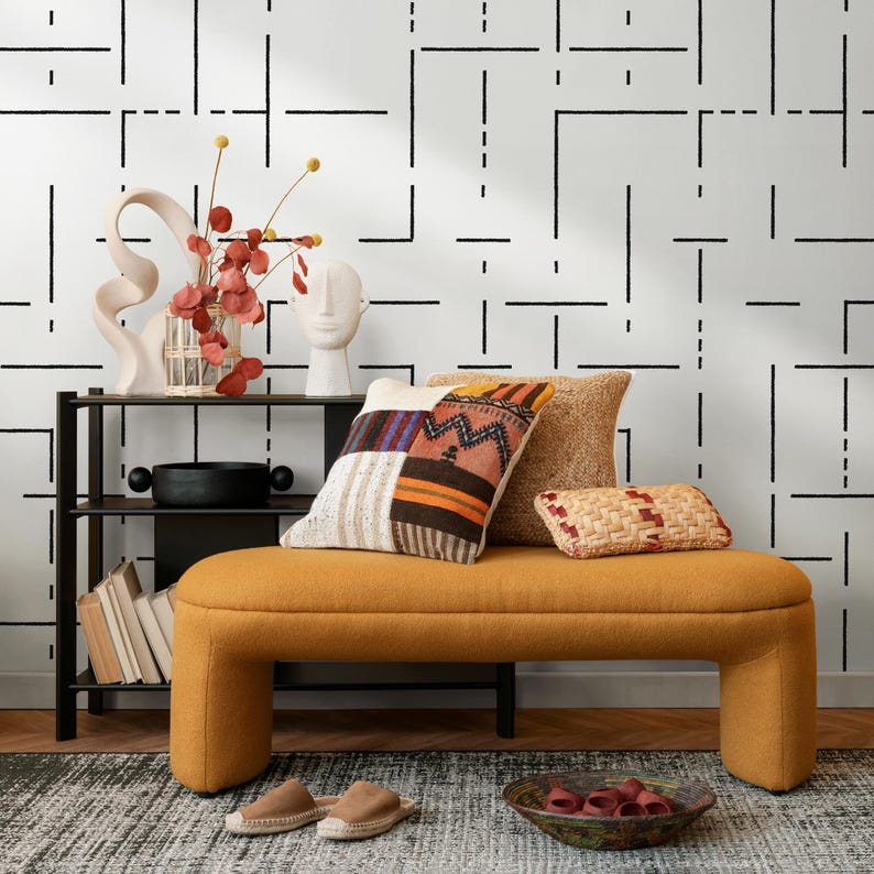 May include: A mustard yellow curved bench with three pillows, one with a colorful patchwork design, one with a natural woven texture, and one with a woven straw design. The bench is in front of a white wall with a black geometric pattern. There is a black shelf with books and a decorative vase with red flowers.