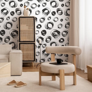 May include: A modern living room with a white and black abstract wallpaper pattern. The room features a light wood chair with a beige cushion, a black mug, and a woven cabinet. The room is decorated in neutral tones.