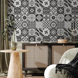 May include: A room interior featuring a black and white patterned tile wall. A wooden cabinet with a woven front sits below the wall. A round wooden side table and a vase with greenery are in the foreground. A chair with a blanket is on the right.
