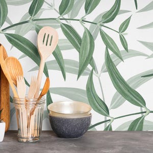 May include: A wooden cutting board, wooden spoons, forks, and a slotted spoon are displayed in a clear glass jar. Two small bowls are stacked, and a small potted succulent sits nearby. The background features a green and white leaf pattern.