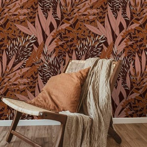 Puede incluir: Una escena interior acogedora con una silla de madera con asiento tejido, un cojín color óxido y una manta de punto trenzado beige. El fondo es un papel pintado con un patrón repetido de hojas estilizadas en tonos marrón, rosa y blanco.