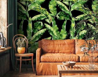 Op de afbeelding: Een woonkamer met een bruine leren bank, houten stoel en salontafel. Het middelpunt is een zwarte muur bedekt met een levendig groen en geel bananenbladpatroon. De kamer heeft een natuurlijk, tropisch thema.