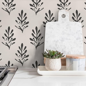 May include: A kitchen setting with a white marble chopping board, a small succulent in a textured pot, and a candle with a wooden lid. The background is wallpaper with a repeating black leaf pattern.