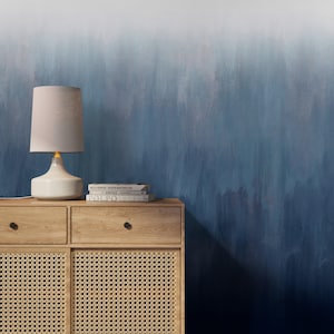 Puede incluir: Una escena interior moderna con un mueble de madera con puertas y cajones de caña tejida. Una lámpara de cerámica blanca con una pantalla beige se encuentra encima del mueble, junto a una pila de libros. El fondo es una pared con gradiente azul.