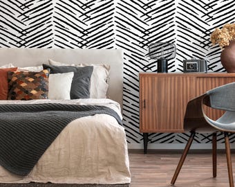 May include: A bedroom with a bed with a white duvet and a black knitted throw blanket. The bed has a white headboard and is against a wall with a black and white geometric pattern wallpaper. There is a wooden dresser with a lamp and a camera on top of it. There is a chair in the corner of the room.