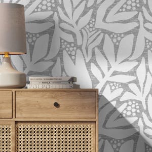 Puede incluir: Un mueble de madera con una puerta de ratán tejido y un cajón. Una lámpara con una pantalla gris se encuentra encima del mueble. El fondo es un papel pintado floral gris y blanco. Los libros están apilados en el mueble con los títulos "Kelly Hoppen Design Masterclass", "Kinfolk" y "Polpo".