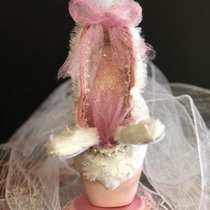 Pode incluir: Uma sapatilha de balé rosa com acabamento em pele branca e um laço de tule rosa. A sapatilha está num suporte rosa com acabamento em renda e está rodeada de tule branco.