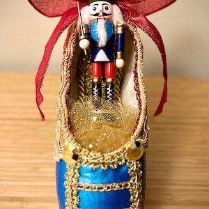 May include: A blue and gold ballet slipper with a nutcracker doll inside. The slipper is decorated with gold trim and rhinestones. A red bow is on top of the slipper.