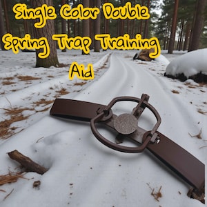 May include: A brown metal spring trap with the text "Single Color Double Spring Trap Training Aid" in yellow. The trap is set in the snow on a path in a forest.