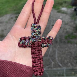 May include: A handmade cross pendant crafted from burgundy and multicolored paracord. The cross features a looped design and a burgundy cord loop for hanging. The intricate weaving creates a textured, durable religious accessory.