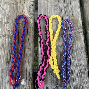 May include: Four braided lanyards in red, blue, black, pink, and yellow, each with a silver-colored key ring. The lanyards are displayed on a weathered wooden surface.
