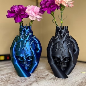 Op de afbeelding: Twee 3D-geprinte hartvormige vazen met schedels en handen die het hart vasthouden. De ene vaas is blauw en de andere is zwart. Beide vazen hebben roze en paarse anjers.