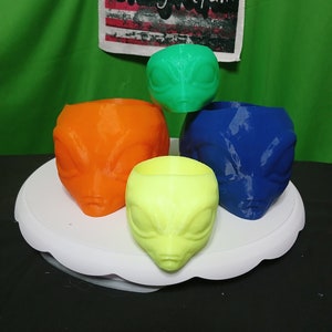 May include: Four alien head planters in different colours: green, orange, blue, and yellow. The planters are arranged on a white pedestal. The text "Crazyvetfam" is visible in the background.