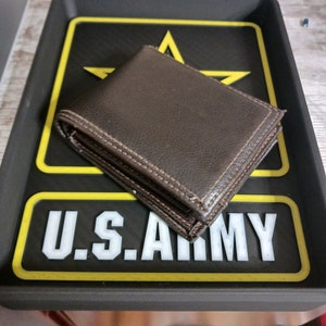 May include: A brown leather wallet with stitched edges rests on a black tray. The tray features a yellow border, a white "U.S. ARMY" text, and a yellow star logo.