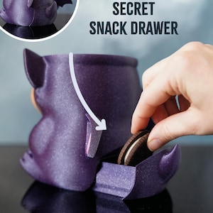 May include: A purple bat-shaped container with a secret snack drawer. The text "SECRET SNACK DRAWER" is above the container. A hand is reaching in to grab a chocolate sandwich biscuit.