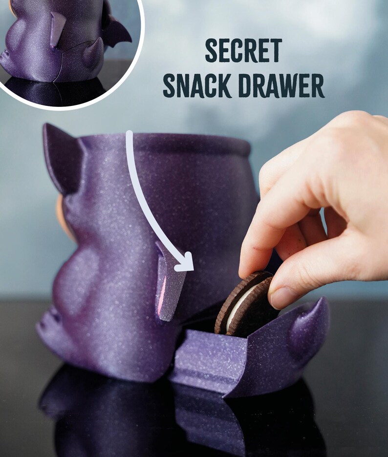 May include: A purple bat-shaped container with a secret snack drawer. The text "SECRET SNACK DRAWER" is above the container. A hand is reaching in to grab a chocolate sandwich biscuit.