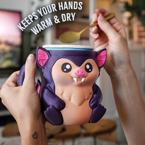 May include: A novelty mug in the shape of a cartoon bat, predominantly purple with pink ears and a tan belly. The mug is held by two hands. The text "KEEPS YOUR HANDS WARM & DRY" is above the mug. A gold spoon is held above the mug.