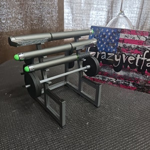 May include: A gray metal pen holder shaped like a weightlifting rack with three pens and a barbell. The rack has a black base and a black barbell with weight plates.