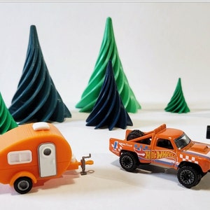 May include: Orange and white toy camper trailer and an orange toy pickup truck with "Hot Wheels" printed on the side. The toys are on a white surface with green and blue 3D printed trees in the background.