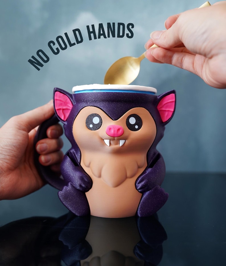 May include: A novelty mug shaped like a cartoon bat, predominantly purple with pink ears and a tan belly. The mug has the text "NO COLD HANDS" above it. A gold spoon is held over the mug.