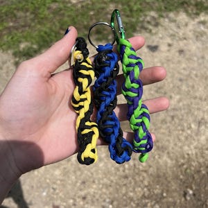 Puede incluir: Tres llaveros trenzados en amarillo y negro, azul y negro, y verde y morado. Cada llavero está unido a un anillo metálico o a un mosquetón. Los llaveros están hechos de un material tejido.